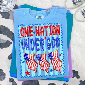 May include: A light blue t-shirt featuring the text ONE NATION UNDER GOD in red and blue letters. Below the text, three American flags hang from a clothesline. The design is framed by a blue scalloped border with small red and blue stars.