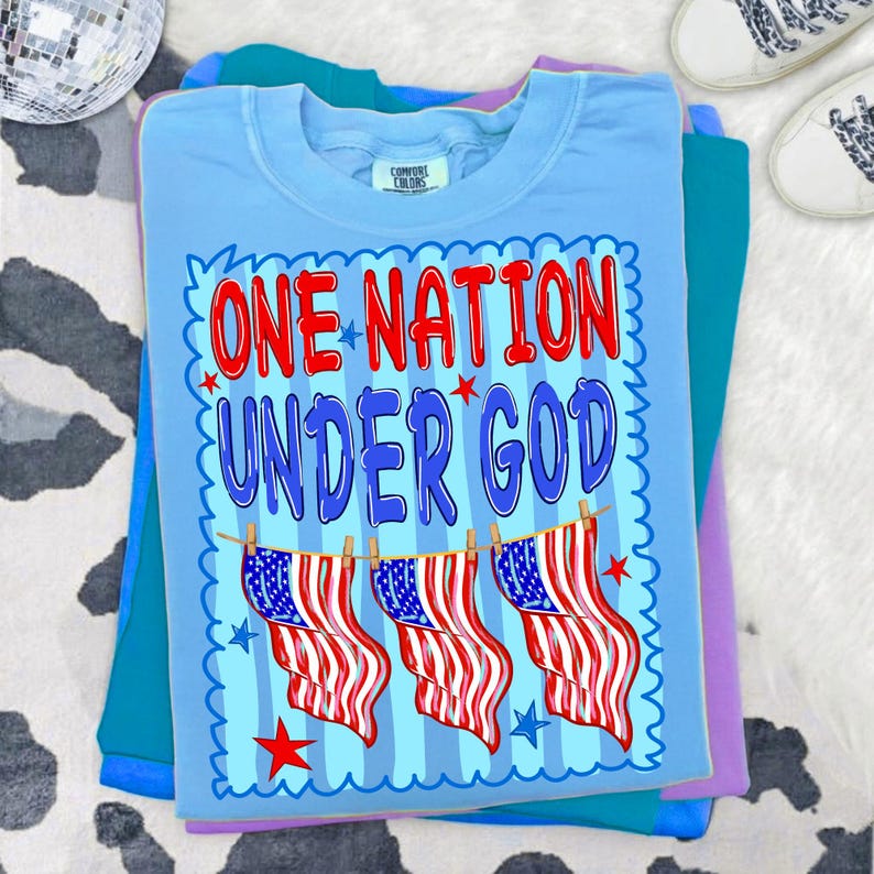 May include: A light blue t-shirt featuring the text ONE NATION UNDER GOD in red and blue letters. Below the text, three American flags hang from a clothesline. The design is framed by a blue scalloped border with small red and blue stars.