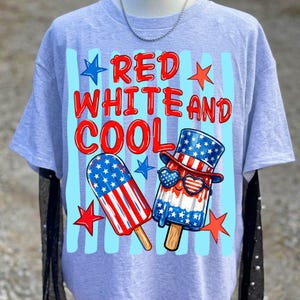 May include: A light gray t-shirt featuring a graphic with the text RED WHITE AND COOL in red letters. The design includes two popsicle illustrations decorated with American flag patterns, one wearing a top hat and heart-shaped sunglasses, with red and blue stars.