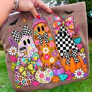 Puede incluir: Una camiseta marr&oacute;n con un gr&aacute;fico de tres fantasmas decorados con estampado de leopardo, patrones florales naranjas y dise&ntilde;os de cuadros blancos y negros, acentuado con calabazas coloridas, margaritas y peque&ntilde;os murci&eacute;lagos.
