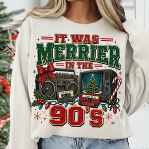 Może przedstawiać: Kremowa bluza z okrągłym dekoltem i nadrukiem graficznym z napisem "IT WAS MERRIER IN THE 90'S". Wz&oacute;r przedstawia zabytkowy radiomagnetofon z czerwoną kokardą, telewizor w stylu retro z choinką oraz dwie kasety magnetofonowe.