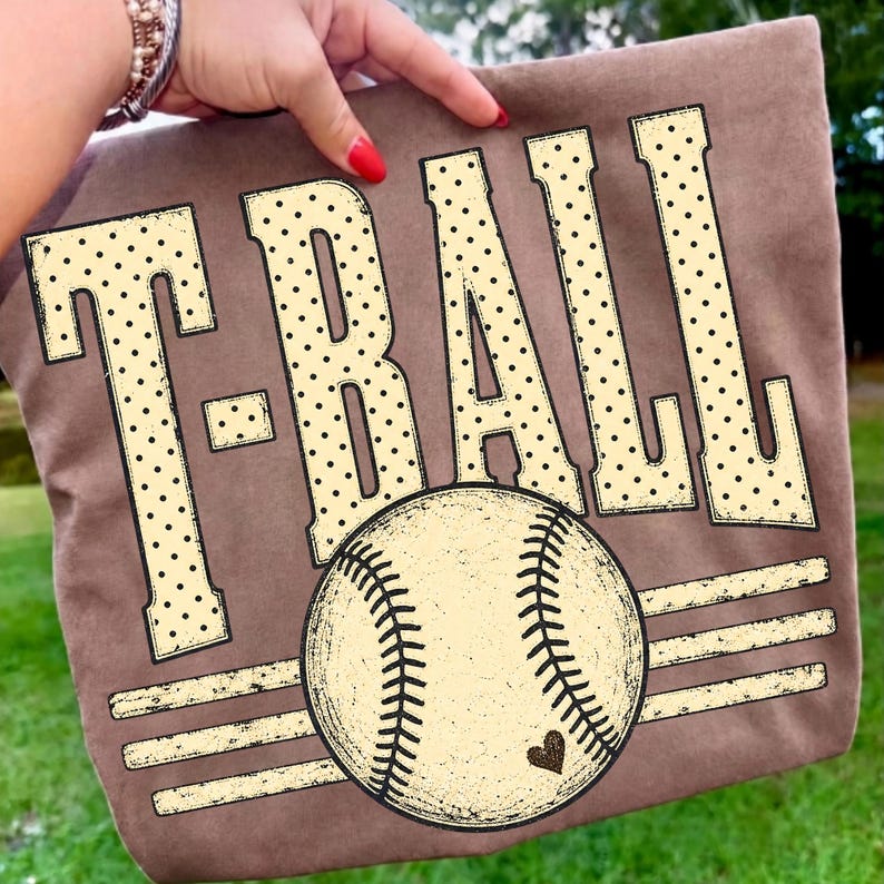 Pode incluir: Uma t-shirt castanha com o texto T-BALL em letras grandes de cor creme com um padr&atilde;o de pontos pretos. Abaixo do texto, encontra-se um gr&aacute;fico de uma bola de basebol com costuras vis&iacute;veis e um pequeno cora&ccedil;&atilde;o, centrado sobre tr&ecirc;s linhas.