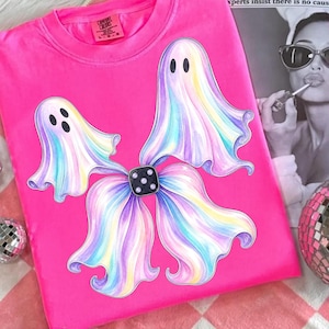May include: A bright pink t-shirt featuring a graphic of two pastel-coloured ghosts with swirling rainbow hues. The ghosts are positioned to form a bow shape, centred by a black square die with white dots. The shirt has a Comfort Colors label.