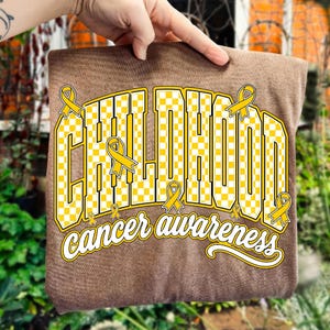 Puede incluir: Una camiseta marr&oacute;n con un dise&ntilde;o gr&aacute;fico que muestra la palabra CHILDHOOD en letras may&uacute;sculas grandes a cuadros amarillos y blancos. El texto cancer awareness aparece debajo en letra cursiva blanca, con cintas amarillas.