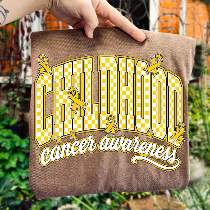 Puede incluir: Una camiseta marr&oacute;n con un dise&ntilde;o gr&aacute;fico que muestra la palabra CHILDHOOD en letras may&uacute;sculas grandes a cuadros amarillos y blancos. El texto cancer awareness aparece debajo en letra cursiva blanca, con cintas amarillas.