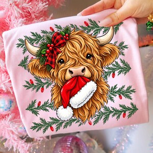 May include: A light pink t-shirt featuring a graphic of a fluffy brown Highland cow with long horns. The cow wears a red and black buffalo plaid bow with holly berries and holds a red and white Santa hat in its mouth, surrounded by pine branches.