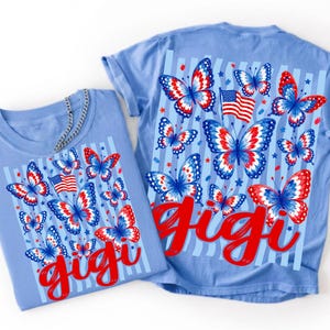 May include: A light blue t-shirt featuring a graphic print of red, white, and blue butterflies, American flags, and vertical stripes. The word "gigi" is written in a large, red, cursive script across the bottom of the butterfly design.