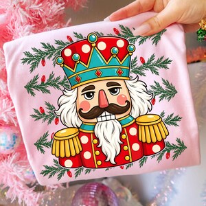Puede incluir: Una camiseta rosa claro con un estampado gr&aacute;fico de un cascanueces que lleva una corona roja y azul verdoso con detalles dorados. El cascanueces tiene un bigote marr&oacute;n, barba blanca y charreteras doradas, rodeado de ramas de pino y luces rojas.