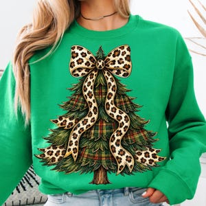 May include: A bright green crewneck sweatshirt featuring a graphic of a Christmas tree with a plaid pattern and a large leopard print bow draped over the top. The tree has detailed green pine needles and a brown trunk, centered on the front of the shirt.