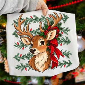 May include: A white t-shirt featuring a cartoon illustration of a brown deer head with large antlers. The deer is adorned with green pine branches, red berries, and a large red ribbon bow. The background pattern consists of green pine needles.