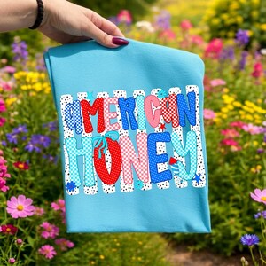 May include: A light blue t-shirt featuring the text AMERICAN HONEY in a bold, multi-colored font. The letters are decorated with red, white, and blue patterns including polka dots, checkers, and stripes, accented by small stars and flowers.