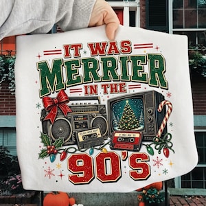 Może przedstawiać: Biały t-shirt z grafiką zawierającą napis IT WAS MERRIER IN THE 90'S. Wz&oacute;r przedstawia zabytkowy radiomagnetofon z czerwoną kokardą, telewizor z choinką, dwie kasety magnetofonowe oraz kolorowe świąteczne lampki choinkowe.
