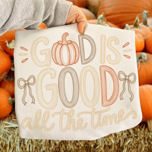May include: A white t-shirt featuring the text "GOD IS GOOD all the time" in a multi-colored, line-art font. The design includes a central pumpkin illustration replacing the letter "O" in "GOD" and two decorative bows on the sides of the text.