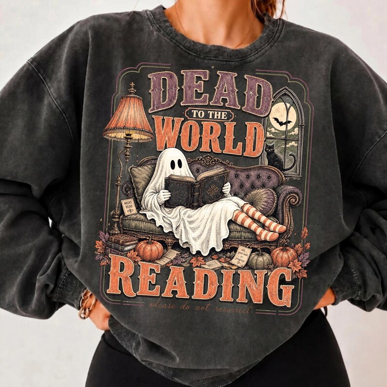 May include: A charcoal gray crewneck sweatshirt featuring a graphic print of a ghost sitting on a sofa reading a book. The text reads DEAD TO THE WORLD READING. The design includes a floor lamp, a window with a moon, a black cat, and pumpkins.