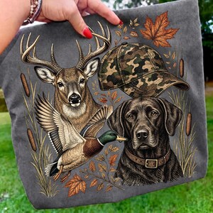 May include: A charcoal gray t-shirt featuring a graphic print of a deer with antlers, a camouflage baseball cap, a flying mallard duck, and a black Labrador retriever wearing a brown collar, surrounded by cattails and orange maple leaves.