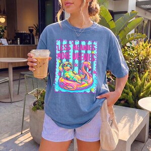 May include: A blue short-sleeve t-shirt featuring a graphic print of a dachshund wearing sunglasses while floating on a pink flamingo pool float. The text "LESS MEANIES MORE WEENIES" is printed in pink above the dog. The shirt is paired with light blue and white striped shorts.