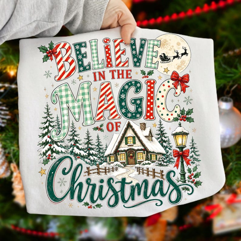 May include: A white t-shirt featuring a colorful graphic with the text "BELIEVE IN THE MAGIC OF CHRISTMAS". The design includes a snowy cabin, pine trees, a street lamp, a moon with a Santa sleigh silhouette, and holly accents in red and green.