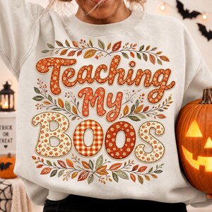 Pu&ograve; includere: Una felpa girocollo grigio chiaro con la scritta "Teaching My Boos" in grandi lettere a motivi arancioni e rossi. Il design &egrave; circondato da un bordo decorativo di foglie autunnali e bacche nei toni dell'arancione, del rosso e del verde.