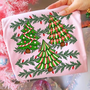 May include: A light pink t-shirt featuring a graphic print of three stylised Christmas trees surrounded by green pine branches and small red light bulbs. One tree has a candy cane stripe pattern, one has a red and green plaid, and one is striped.