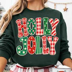 May include: A dark green long-sleeve sweatshirt featuring the text HOLLY JOLLY in large, multi-patterned letters. The letters include red bows, green polka dots, red and white plaid, green Christmas trees, red stars, and red stripes.