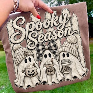 May include: A brown fabric item featuring an embroidered design of three ghosts with the text Spooky Season. The ghosts wear knit hats and a plaid bow, holding a pumpkin mug and coffee cups with maple leaf details against a fence background.