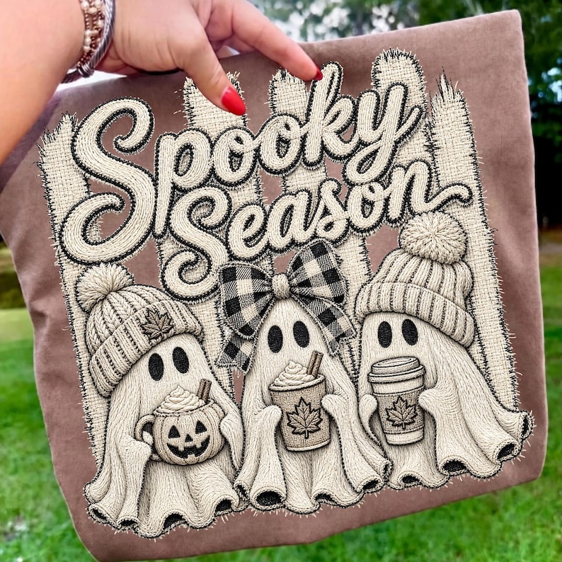 May include: A brown fabric item featuring an embroidered design of three ghosts with the text Spooky Season. The ghosts wear knit hats and a plaid bow, holding a pumpkin mug and coffee cups with maple leaf details against a fence background.
