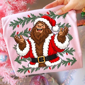 Puede incluir: Una camiseta rosa claro con un gr&aacute;fico de una criatura marr&oacute;n y peluda que lleva un traje de Pap&aacute; Noel rojo con ribetes de piel blanca, un cintur&oacute;n negro con hebilla dorada y un gorro rojo, rodeada de ramas de pino verdes y luces rojas.