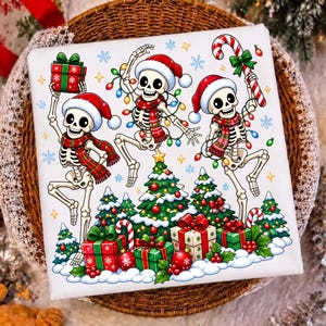 Op de afbeelding: Een wit vierkant decoratie-item met een feestelijke illustratie van drie dansende skeletten met kerstmutsen en geruite sjaals. De skeletten worden omringd door kerstbomen, ingepakte cadeaus, kerstverlichting en een zuurstok.