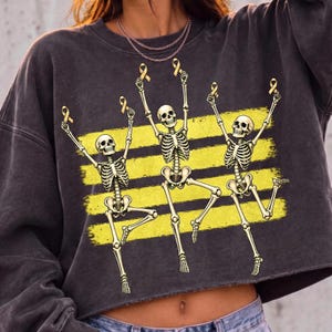 May include: A charcoal grey long-sleeve sweatshirt featuring a graphic print of three dancing skeletons. Each skeleton holds a small yellow awareness ribbon above its head. The design is set against three horizontal yellow brushstroke stripes.