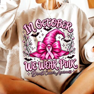 May include: A beige sweatshirt featuring a graphic print of a bright pink witch hat adorned with a large bow and a breast cancer awareness ribbon. The text reads In October We Wear Pink Breast Cancer Awareness, surrounded by small black bats.