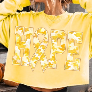 May include: A bright yellow crewneck sweatshirt featuring the word HOPE printed in large block letters across the chest. The letter O is replaced by an awareness ribbon. The letters have a yellow and white camouflage pattern with a distressed texture.