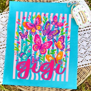 以下が含まれることがあります： 鮮やかなターコイズブルーのTシャツ。ピンク、紫、オレンジ、ティールカラーのカラフルな蝶のグラフィックプリントが、垂直のピンクのストライプの背景に描かれています。中央には「gigi」という文字が、大きく太いホットピンクの筆記体でプリントされています。