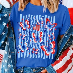 Puede incluir: Una camiseta azul real con un estampado gr&aacute;fico de varias mariposas con patrones de alas en rojo, blanco y azul. El dise&ntilde;o incluye una peque&ntilde;a bandera estadounidense y rayas verticales azul claro en el fondo, acentuadas por peque&ntilde;as estrellas rojas.