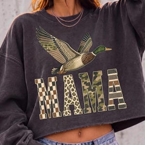May include: A charcoal gray cropped sweatshirt featuring a graphic of a flying mallard duck above the word MAMA. The letters are filled with various patterns including a brown and white checkerboard, leopard print, and green camouflage designs.
