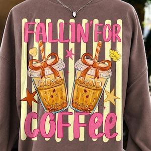 K&ouml;nnte beinhalten: Ein braunes Sweatshirt mit Rundhalsausschnitt und einem Grafikdruck von zwei Eiskaffeebechern mit orangefarbenen Schleifen und Strohhalmen. Der Text FALLIN' FOR COFFEE in Pink auf vertikalen cremefarbenen Streifen, verziert mit kleinen Sternen und Bl&auml;ttern.