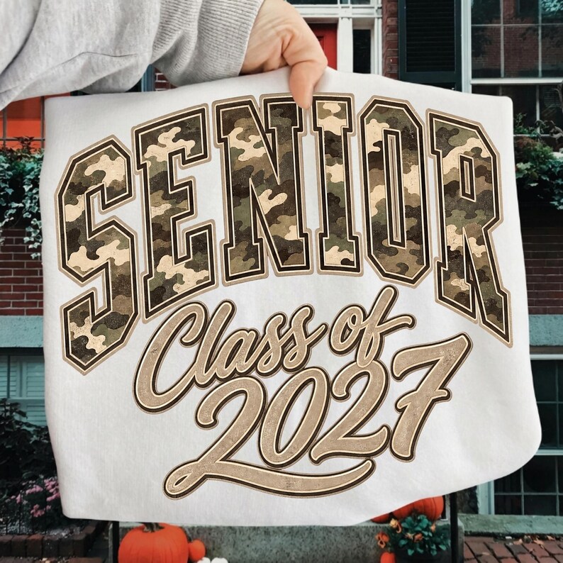 May include: A white sweatshirt featuring the word SENIOR in large, block-style letters with a green, brown, and tan camouflage pattern. Below, the text Class of 2027 is printed in a large, tan, cursive script with a dark brown outline.