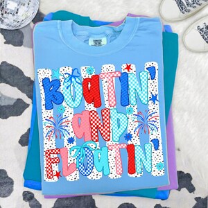Op de afbeelding: Een lichtblauw t-shirt met de tekst "BOATIN' AND FLOATIN'" in een veelkleurig, gedessineerd lettertype. Het ontwerp bevat rood, blauw en wit geblokte en gestippelde letters, kleine stervormen en vuurwerkafbeeldingen op een witte achtergrond.