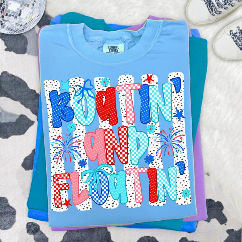 Op de afbeelding: Een lichtblauw t-shirt met de tekst "BOATIN' AND FLOATIN'" in een veelkleurig, gedessineerd lettertype. Het ontwerp bevat rood, blauw en wit geblokte en gestippelde letters, kleine stervormen en vuurwerkafbeeldingen op een witte achtergrond.
