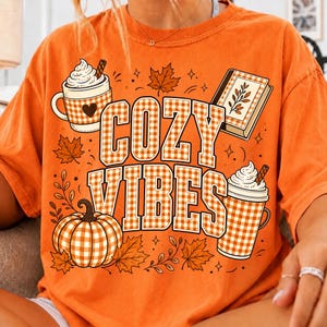 Puede incluir: Una camiseta naranja con un estampado gr&aacute;fico que incluye el texto COZY VIBES en un patr&oacute;n de cuadros vichy blanco y naranja. El dise&ntilde;o muestra una taza de chocolate caliente, un libro, una calabaza y hojas de arce con cuadros a juego.