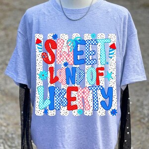 K&ouml;nnte beinhalten: Ein hellgraues T-Shirt mit einem Grafikdruck, der den Text SWEET LAND OF LIBERTY in roten, wei&szlig;en und blauen Mustern zeigt. Das Design umfasst vertikale gepunktete Streifen, kleine Blumen, Sterne und Raketensymbole in patriotischen Farben.