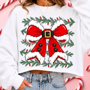 Puede incluir: Una sudadera corta blanca con un gr&aacute;fico grande de un lazo rojo estilo Santa Claus con borde de piel blanca, un cintur&oacute;n negro con hebilla dorada y cuatro botones negros sobre un fondo de ramas de pino verdes y luces rojas.