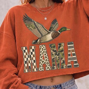 May include: A burnt orange long-sleeve sweatshirt featuring a graphic of a flying mallard duck above the word MAMA. The letters are filled with different patterns including a brown checkerboard, leopard print, green camouflage, and horizontal stripes.