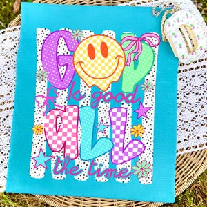 May include: A bright turquoise t-shirt featuring a graphic print with the text "GOD is good ALL the time". The design includes a yellow checkered smiley face, colorful polka-dot and checkered letters, pink bows, small stars, and various flower icons.