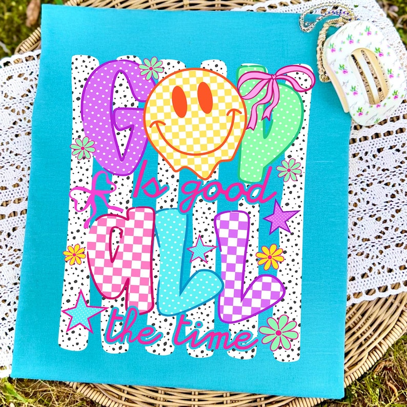 May include: A bright turquoise t-shirt featuring a graphic print with the text "GOD is good ALL the time". The design includes a yellow checkered smiley face, colorful polka-dot and checkered letters, pink bows, small stars, and various flower icons.