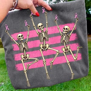 May include: A dark gray tote bag featuring a graphic print of three beige skeletons in dynamic poses, each holding a pink breast cancer awareness ribbon above their heads, set against a background of three horizontal pink brushstroke stripes.