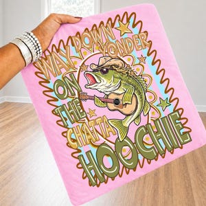 May include: Pink blanket with a graphic of a bass playing a guitar, wearing sunglasses and a cowboy hat. Text reads "Way Down Yonder On The Chattahoochee". The blanket is held by a person wearing silver bracelets.