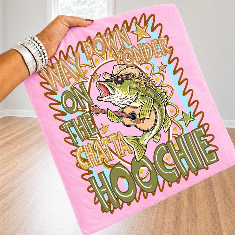 May include: Pink blanket with a graphic of a bass playing a guitar, wearing sunglasses and a cowboy hat. Text reads "Way Down Yonder On The Chattahoochee". The blanket is held by a person wearing silver bracelets.