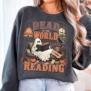 May include: A charcoal gray sweatshirt featuring a graphic print of a ghost sitting on a sofa reading a book. The text reads DEAD TO THE WORLD READING. The illustration includes a lamp, a black cat, a window with a bat, pumpkins, and autumn leaves.