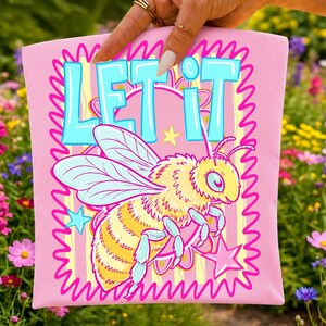 Peut inclure: Un carr&eacute; de tissu rose clair avec une illustration graphique d'une abeille jaune et bleu clair. Le texte "LET IT" est imprim&eacute; en grandes lettres bleu clair en haut. Le motif comprend un fond ray&eacute; jaune, des &eacute;toiles roses et une bordure rose ondul&eacute;e.