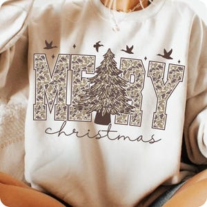 May include: Cream-colored sweatshirt with the words "MERRY" in large, camouflage-patterned letters, a detailed Christmas tree, and the word "christmas" in script. The design includes bird and star accents.